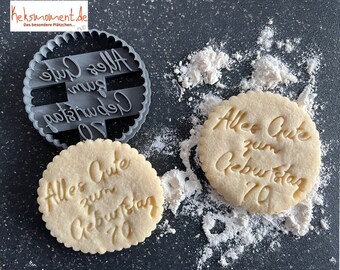 May include: A close-up of two cookie cutters, one with the text "Alles Gute zum Geburtstag 70" and the other with a cookie already cut out with the same text. The cookie cutters are on a black surface with white powder.