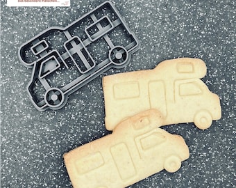 May include: A gray plastic camper cookie cutter next to two baked camper-shaped cookies. The cookies are golden brown with detailed impressions from the cutter. The cookie cutter has the text "keks moment.de" in the upper left corner.