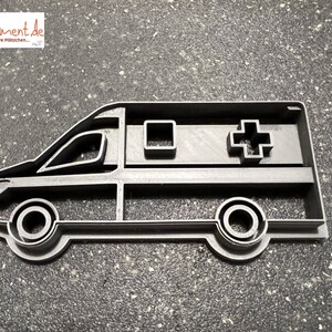 May include: A black plastic cookie cutter shaped like an ambulance. The ambulance has a cross on the side and a window on the side.