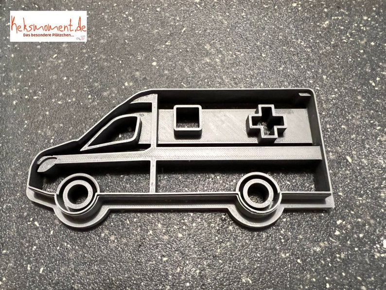 May include: A black plastic cookie cutter shaped like an ambulance. The ambulance has a cross on the side and a window on the side.