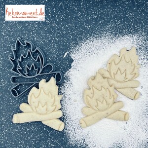 Puede incluir: Un cortador de galletas de plástico gris en forma de fogata con dos troncos y llamas. Se muestran tres galletas, cada una con el mismo diseño, sobre una superficie blanca.