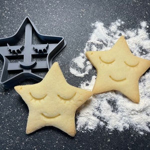 Può includere: Due biscotti a forma di stella con gli occhi chiusi e una bocca sorridente, accanto a uno stampino per biscotti a forma di stella in plastica nera con lo stesso design.