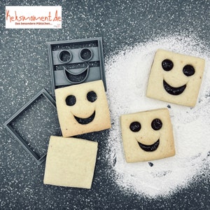 Può includere: Tre biscotti quadrati con un designo a forma di viso sorridente. I biscotti sono decorati con una farcitura marrone scuro. I biscotti sono su una superficie grigia con zucchero a velo bianco.