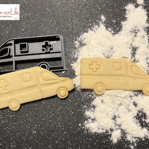 May include: A black plastic cookie cutter shaped like an ambulance, with a red cross on the side, and a light brown cookie in the shape of an ambulance next to it.