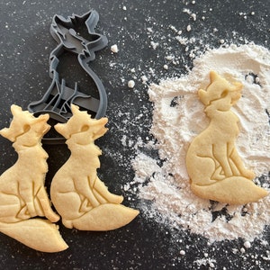 Puede incluir: Tres galletas con forma de zorro, dos están sobre una superficie negra y una sobre una superficie blanca. Las galletas son de color marrón claro y tienen un diseño de zorro detallado. Un cortador de galletas con forma de zorro gris está en el fondo.