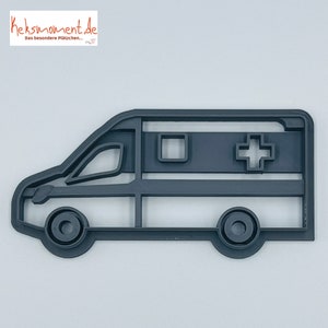 May include: A grey plastic cookie cutter shaped like an ambulance. The ambulance has a white cross on the side and a square window.