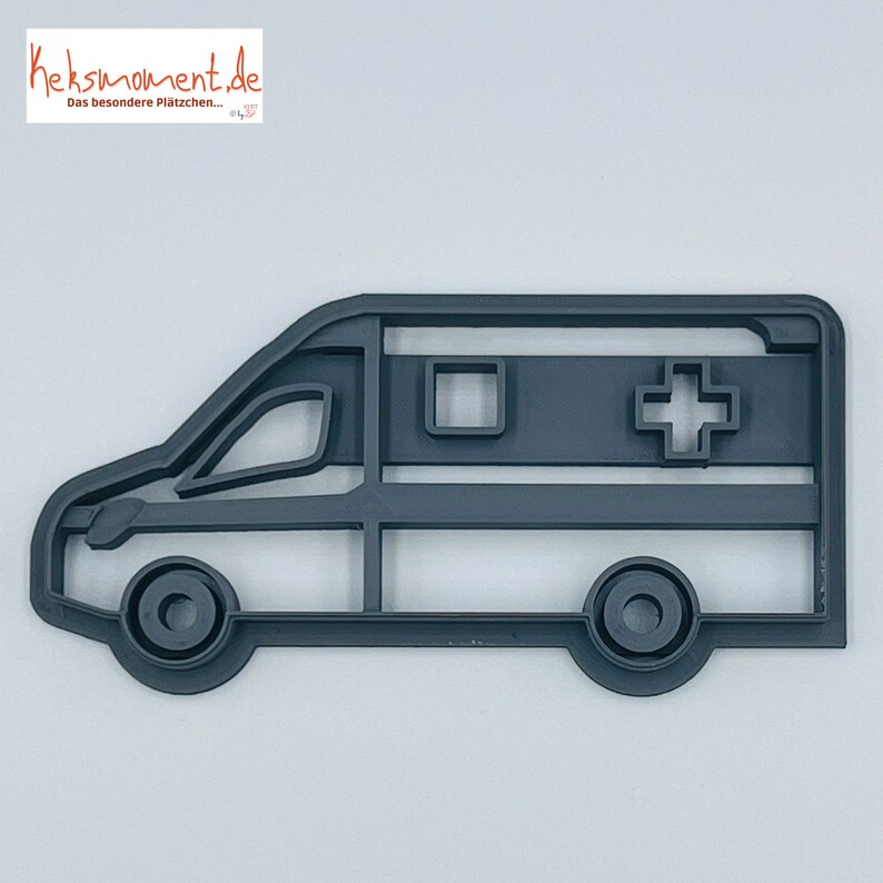 May include: A grey plastic cookie cutter shaped like an ambulance. The ambulance has a white cross on the side and a square window.