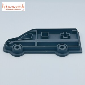 May include: A grey plastic cookie cutter shaped like an ambulance. The cutter has a red cross on the side and a window on the front.