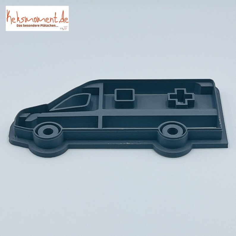 May include: A grey plastic cookie cutter shaped like an ambulance. The cutter has a red cross on the side and a window on the front.