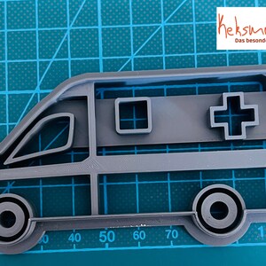 May include: Grey plastic cookie cutter shaped like an ambulance. The ambulance has a red cross on the side and a small window.