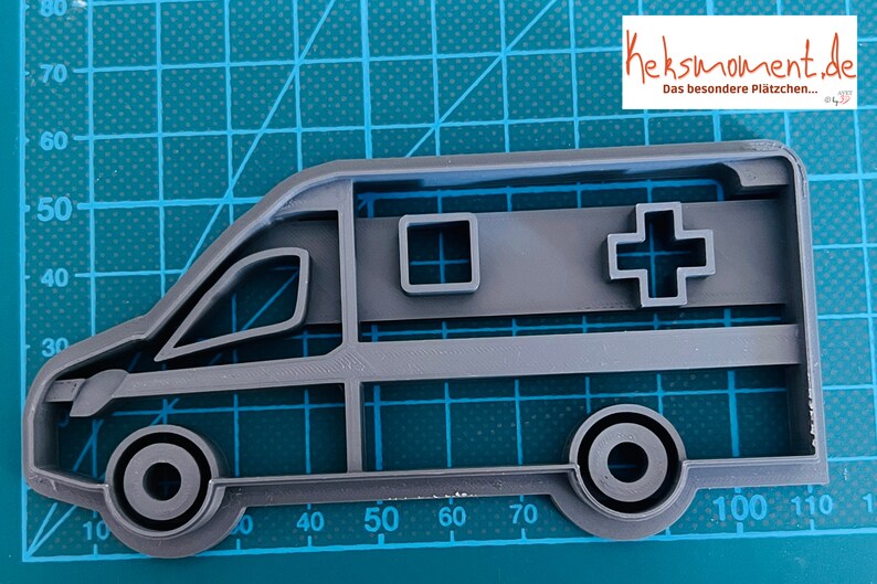 May include: Grey plastic cookie cutter shaped like an ambulance. The ambulance has a red cross on the side and a small window.