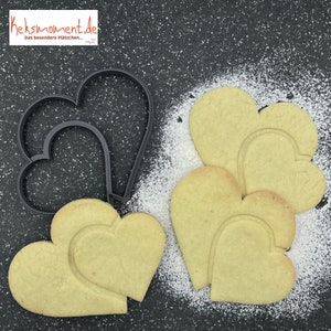 Puede incluir: Tres galletas con forma de corazón, dos apiladas una encima de la otra, con un cortador de galletas con forma de corazón sobre una superficie negra. Las galletas son de color marrón claro y el cortador de galletas es negro.