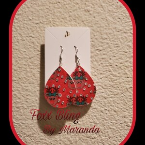 May include: Red teardrop earrings with a reindeer pattern and silver hooks. The earrings feature a repeating pattern of reindeer faces with teal bows and silver accents. The text "Foxx Bling By Maranda" is visible at the bottom.