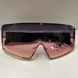 May include: A pair of oversized sunglasses with a black frame and pink and purple gradient lenses. The sunglasses have gold accents and a CE mark on the right lens.