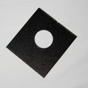 Puede incluir: Una pieza cuadrada de material negro con un agujero redondo en el centro. El material tiene una superficie texturizada.