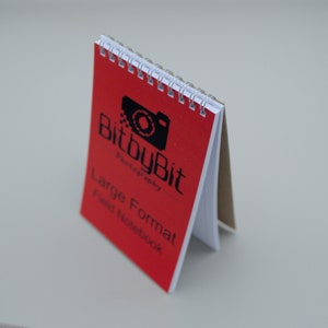 May include: A red spiral-bound field notebook with the text "BitbyBit Photography" and "Large Format Field Notebook" printed in black. The notebook is open, revealing white and tan pages.