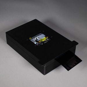 May include: A black plastic slide scanner with a clear window to view the slide. A slide is partially inserted into the scanner.