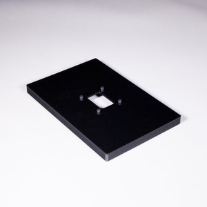 Może przedstawiać: Czarna plastikowa płytka z prostokątnym wycięciem i czterema małymi otworami na śruby.