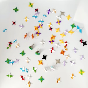 May include: An assortment of colorful origami star earrings and individual stars scattered on a white surface. The earrings and stars are in various colors, including green, yellow, orange, red, purple, and blue. Some earrings are stud style.