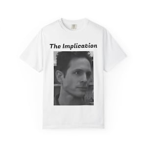 May include: White t-shirt featuring a black and white image of a man's face, with the text "The Implication" above the image. The shirt is a crew neck style.