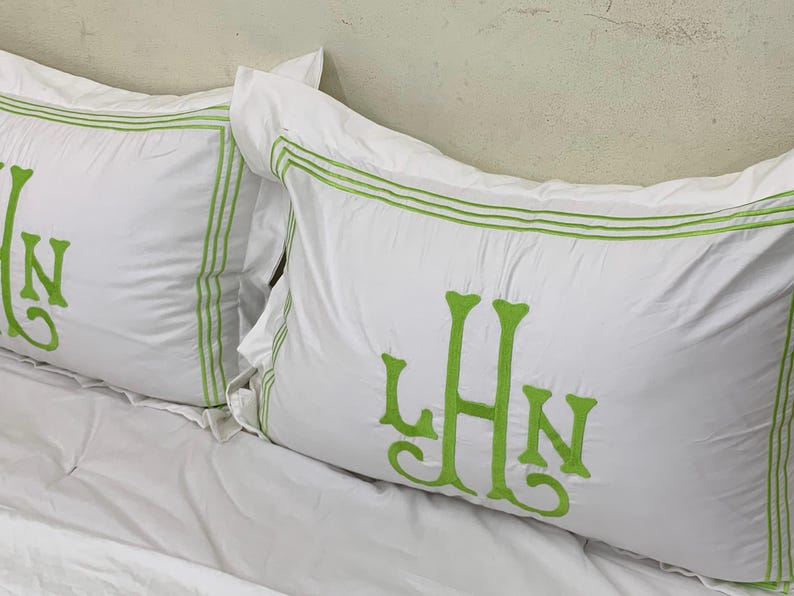 Puede incluir: Dos fundas de almohada rectangulares blancas con iniciales bordadas en verde y ribete verde. Las iniciales "LHN" se muestran en la parte delantera de cada almohada. Las fundas de almohada est&aacute;n sobre una s&aacute;bana blanca.