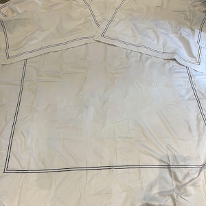 May include: White bedding with a black double border around the edges. The bedding is folded in half and laid flat on a gray surface.