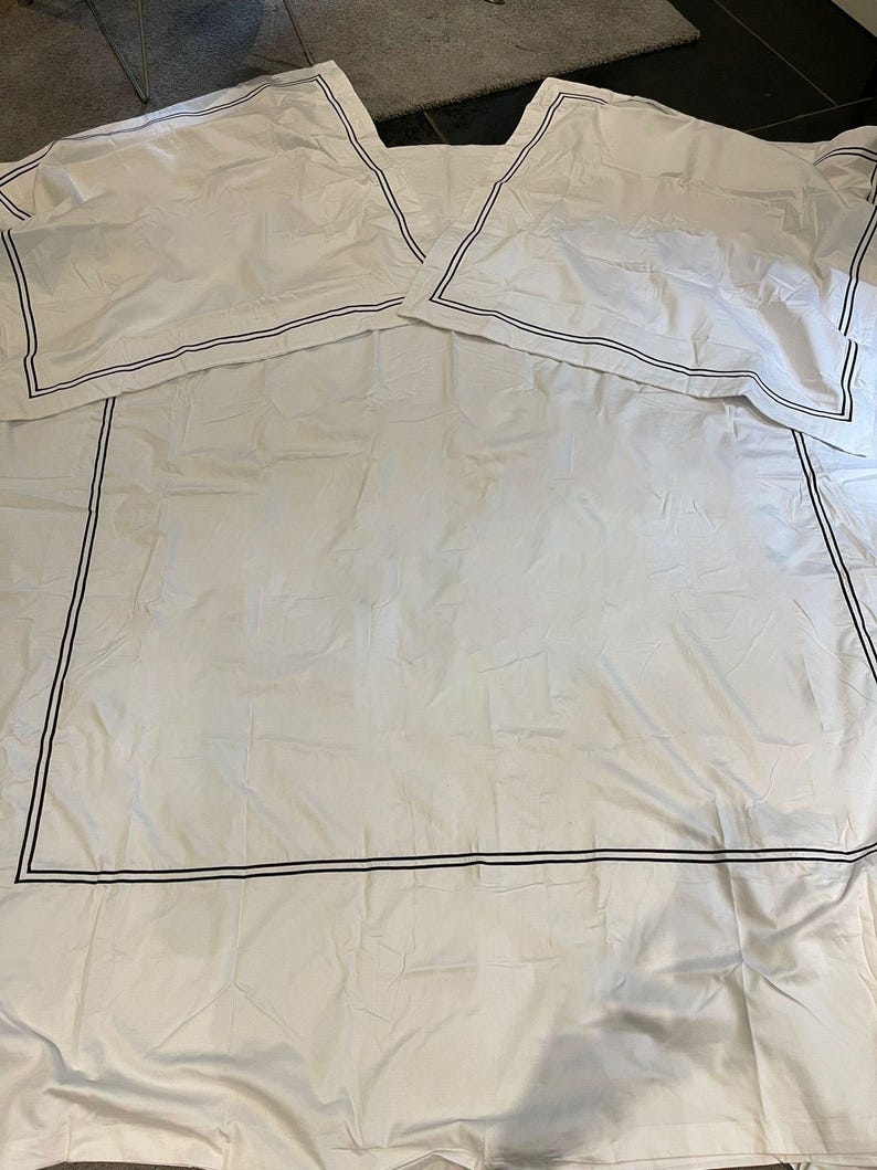 May include: White bedding with a black double border around the edges. The bedding is folded in half and laid flat on a gray surface.