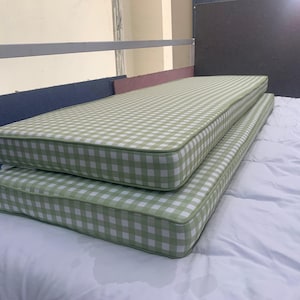 Puede incluir: Dos cojines rectangulares con estampado de cuadros vichy verdes y blancos. Los cojines están apilados, con un borde blanco y un ribete verde. Se encuentran sobre un edredón blanco. Los cojines miden unos 150 cm de largo.
