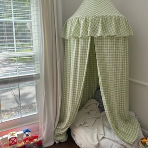 May include: A green and white gingham canopy bed with a ruffled edge. The canopy is set up in a corner with a window and white curtains. A white mattress and blankets are inside the canopy.