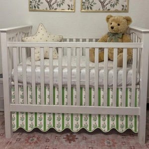 Puede incluir: Una cuna de madera blanca con un faldón festoneado de rayas verticales verdes y blancas con pequeños motivos florales. Dentro de la cuna hay un colchón blanco con un patrón claro, un cojín en forma de estrella y un oso de peluche beige.