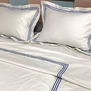 Pode incluir: Len&ccedil;&oacute;is de cama brancos com listras bordadas azuis. Os len&ccedil;&oacute;is est&atilde;o dobrados e dispostos em uma cama com duas almofadas brancas com listras bordadas azuis.