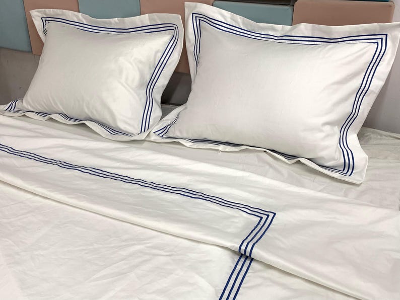 Pode incluir: Len&ccedil;&oacute;is de cama brancos com listras bordadas azuis. Os len&ccedil;&oacute;is est&atilde;o dobrados e dispostos em uma cama com duas almofadas brancas com listras bordadas azuis.