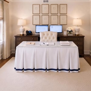 May include: A white desk with a glass top and a pleated skirt featuring a navy blue border trim. The desk is positioned in front of a dark wood credenza with two white lamps, two computer monitors, and a tufted white office chair in the center.
