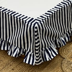 May include: A navy and white striped bed skirt with a frilled edge. The fabric features vertical stripes and is draped over a bed corner. The bed skirt is on a brown woven rug.
