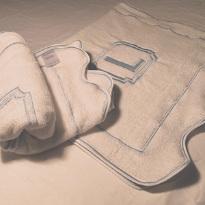 May include: A white bath mat with a blue embroidered border and a blue embroidered letter "L" in the center. A white towel is rolled up and placed on top of the mat.