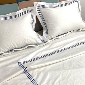 Pode incluir: Len&ccedil;&oacute;is de cama brancos com uma borda bordada azul. Os len&ccedil;&oacute;is est&atilde;o dobrados e dispostos em uma cama com dois travesseiros brancos com bordas bordadas azuis.
