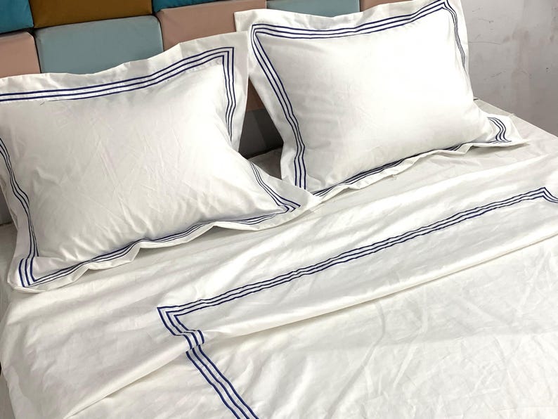 Pode incluir: Len&ccedil;&oacute;is de cama brancos com uma borda bordada azul. Os len&ccedil;&oacute;is est&atilde;o dobrados e dispostos em uma cama com dois travesseiros brancos com bordas bordadas azuis.