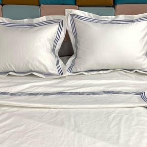 Pode incluir: Len&ccedil;&oacute;is de cama brancos com uma borda bordada azul. Os len&ccedil;&oacute;is est&atilde;o dobrados e dispostos em uma cama com duas almofadas brancas com bordas bordadas azuis.