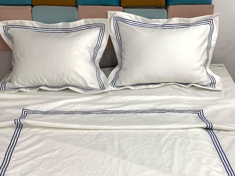 Pode incluir: Len&ccedil;&oacute;is de cama brancos com uma borda bordada azul. Os len&ccedil;&oacute;is est&atilde;o dobrados e dispostos em uma cama com duas almofadas brancas com bordas bordadas azuis.