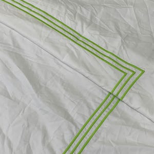 Puede incluir: Tejido blanco con una textura sutil, con tres l&iacute;neas paralelas de color verde lima que forman un patr&oacute;n geom&eacute;trico. Las l&iacute;neas est&aacute;n cosidas, creando un borde decorativo. La tela parece ligeramente arrugada.