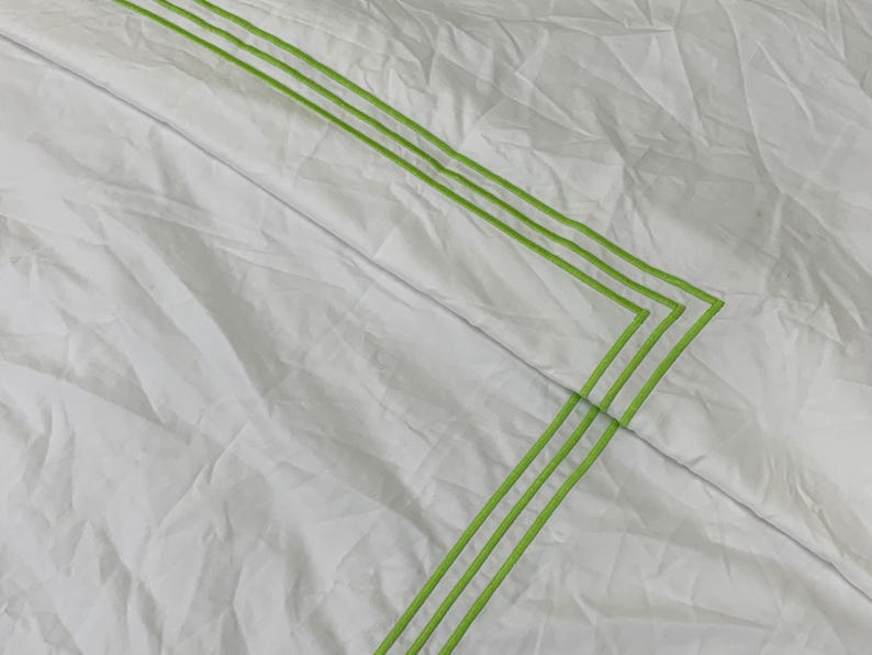 Puede incluir: Tejido blanco con una textura sutil, con tres l&iacute;neas paralelas de color verde lima que forman un patr&oacute;n geom&eacute;trico. Las l&iacute;neas est&aacute;n cosidas, creando un borde decorativo. La tela parece ligeramente arrugada.