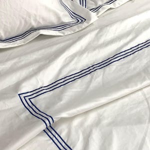 Pode incluir: Len&ccedil;&oacute;is de cama brancos com uma borda azul. A borda &eacute; uma s&eacute;rie de linhas paralelas.