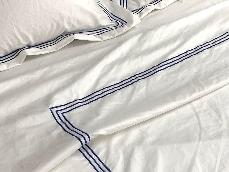Pode incluir: Len&ccedil;&oacute;is de cama brancos com uma borda azul. A borda &eacute; uma s&eacute;rie de linhas paralelas.