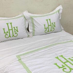 Puede incluir: Juego de cama blanco con ribete verde e iniciales bordadas. El juego incluye dos fundas de almohada y una funda n&oacute;rdica, todas con un monograma en color verde vibrante. La ropa de cama est&aacute; cuidadosamente colocada sobre una cama.
