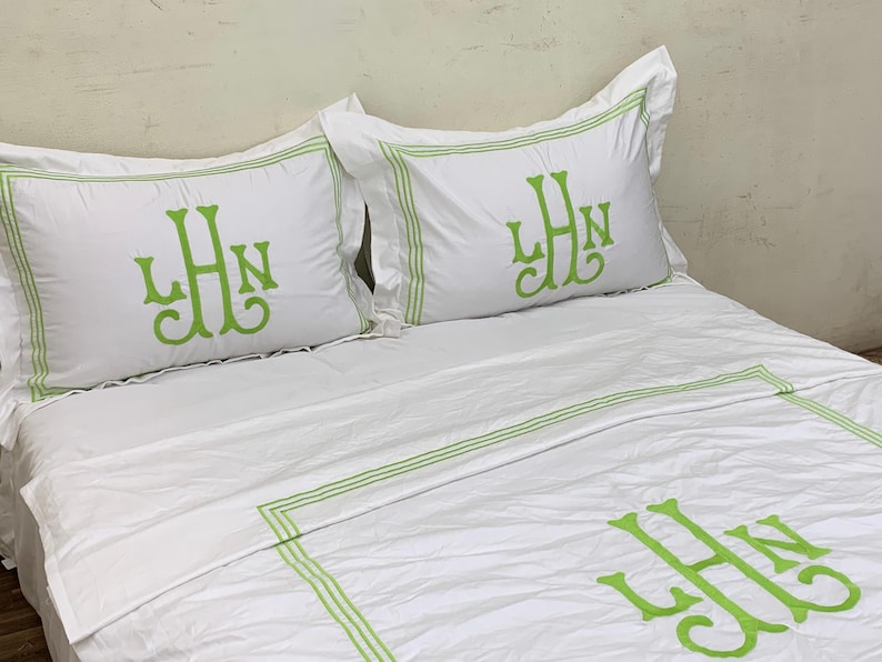 Puede incluir: Juego de cama blanco con ribete verde e iniciales bordadas. El juego incluye dos fundas de almohada y una funda n&oacute;rdica, todas con un monograma en color verde vibrante. La ropa de cama est&aacute; cuidadosamente colocada sobre una cama.