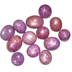 Puede incluir: Colección de gemas de rubí estrella en varias formas, incluyendo ovaladas y redondas. Las gemas muestran una gama de tonos rosados y púrpuras, con un efecto de estrella visible en algunas. Sobre un fondo blanco.