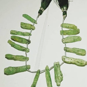 May include: A strand of green kyanite gemstone beads. The beads are oblong and vary in size and shape, strung on a silver-colored wire. The ends of the strand are finished with dark green tassels and silver-colored accents. The kyanite has a natural, textured appearance.