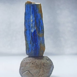 Pu&ograve; includere: Un cristallo blu e oro si trova in cima a una roccia grigia. Il cristallo &egrave; lungo e sottile, con una punta appuntita. La roccia &egrave; rotonda e liscia.