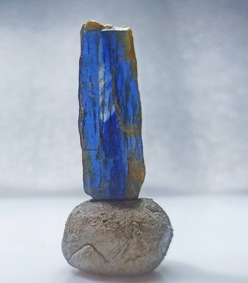 Pu&ograve; includere: Un cristallo blu e oro si trova in cima a una roccia grigia. Il cristallo &egrave; lungo e sottile, con una punta appuntita. La roccia &egrave; rotonda e liscia.