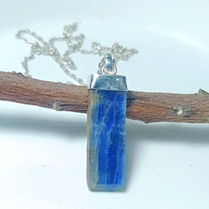 May include: A rectangular blue gemstone pendant with a silver-coloured metal cap and chain. The gemstone has a textured surface and is suspended on a delicate silver chain. The pendant is displayed on a brown wooden branch.